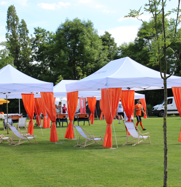 Serie van witte 6x3 m evenementen gazebos met oranje hoekgordijnen gepersonaliseerd met IngDiba logo en witte druppelvlaggen voor een buitenevenement op een groot sportveld