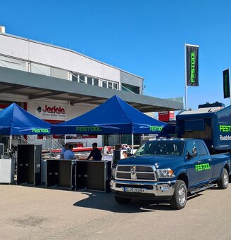 3x3m blaues Eventzelt mit grünem Festool-Logo kombiniert mit Firmenwagen und Wohnwagen vor dem Baumarkt Jedele