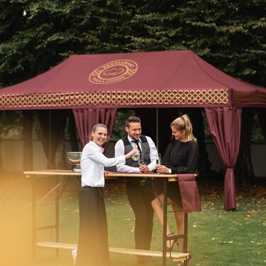 Eleganter Faltpavillon 8x4 m in Burgunderrot mit goldenen Verzierungen und dekorativen Vorhängen, die eine Stange bedecken. Der Pavillon steht auf einer Wiese und wird für den Empfang einer eleganten Veranstaltung verwendet, während die Leute Sekt von einer Kellnerin serviert trinken