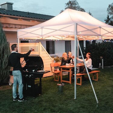 Freunde feiern unter dem Gartenzelt eine Grill- und Gartenparty. Das Gartenzelt ist feierlich dekoriert mit Lichterketten.