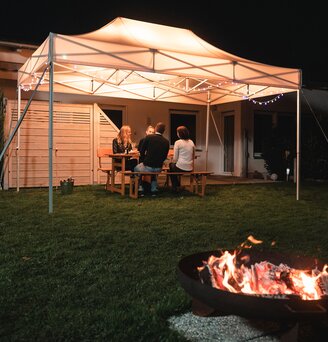 Das 4,5x3 m Gartenzelt steht im Dunkeln im Garten. Darunter sitzen die 4 Gäste. Vor dem Zelt ist noch eine Feuerstelle.