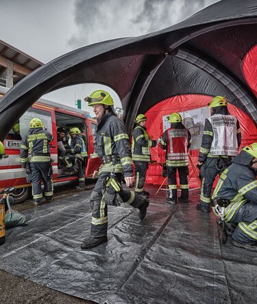 Feuerwehrkräfte bei der Arbeit in und vor einem rot-schwarzen pneumatischen Einsatzzelt neben einem Einsatzfahrzeug. 